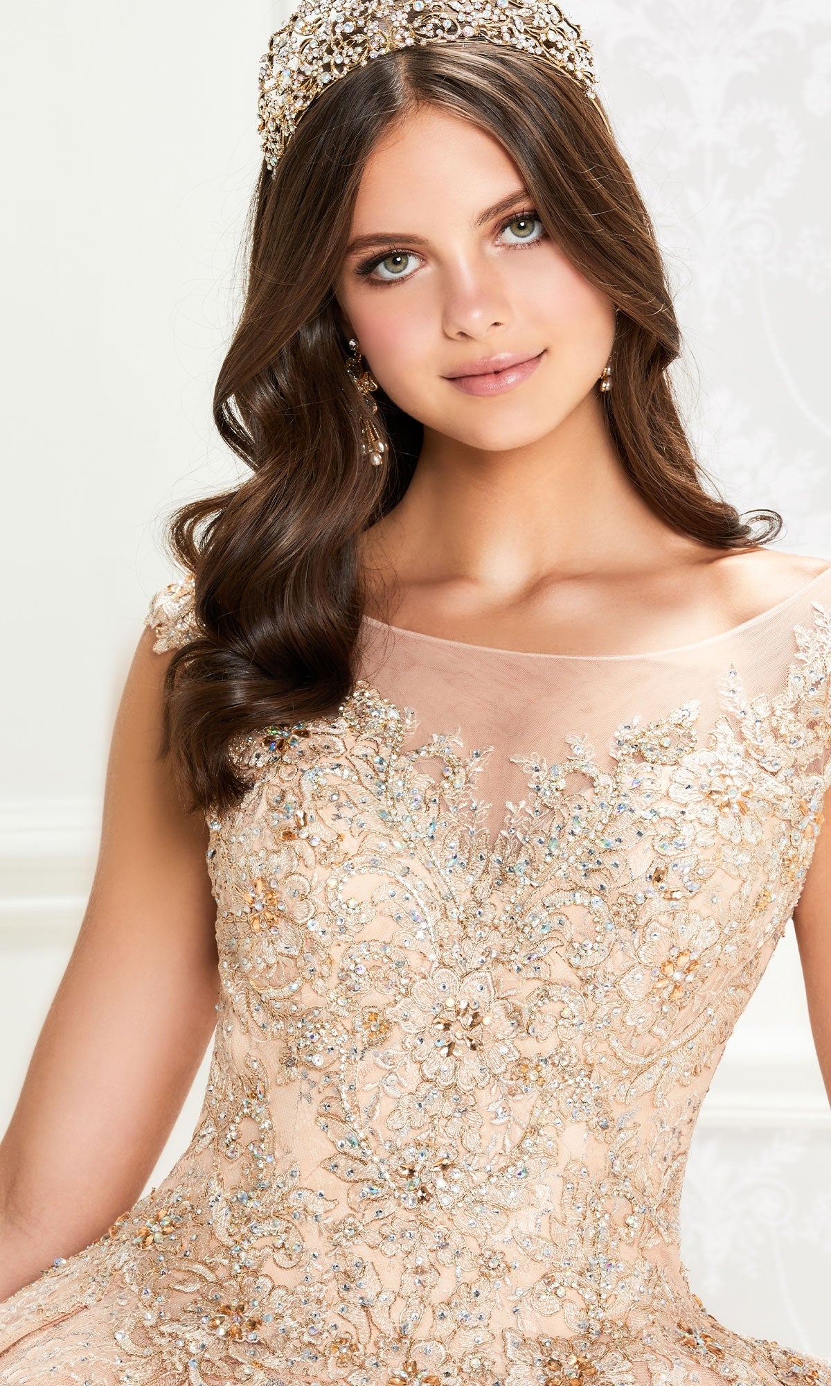 A woman poses indoors against a light background wearing the Princesa Traditional Lace Quinceañera Dress PR12002, featuring an ornate beaded bodice, sheer neckline, floral embroidery, and accessorized with a jeweled headpiece and earrings.